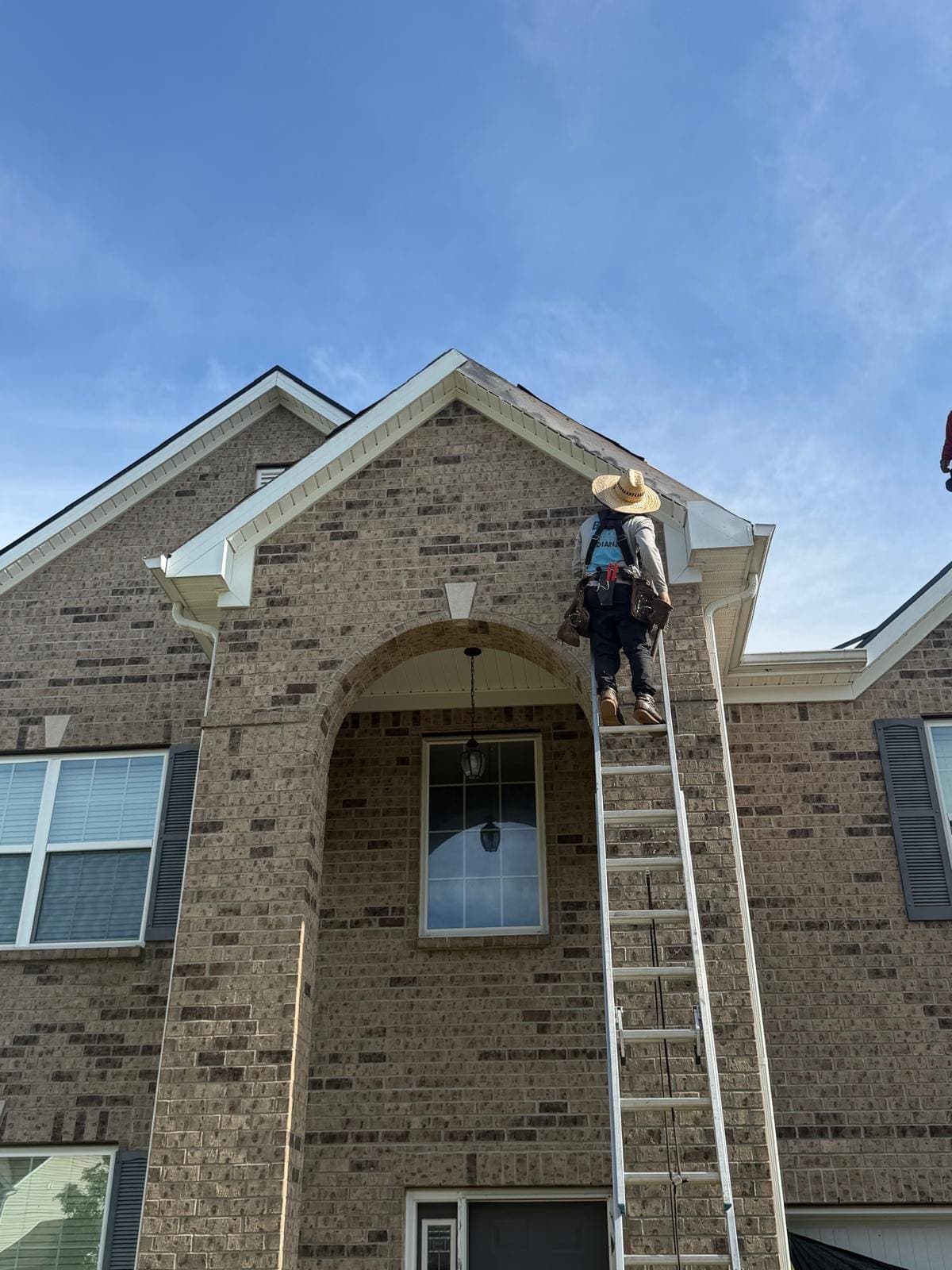 Roofer in a sun hat and tool belt climbing the ladder to the roof of a brick Avon home